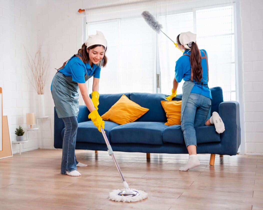 One woman is mopping the wooden floor, while the other is dusting near a window behind a blue sofa with yellow cushions